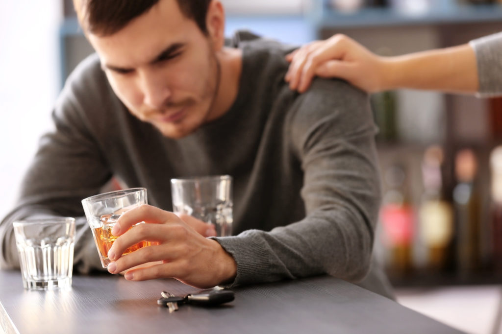 Na zdjęciu widoczny mężczyzna siedzący przy stole, który w ręku trzyma szklankę z alkoholem. Na jego ramieniu spoczywa w geście wsparcia kobieca dłoń. Kadr amerykański. 