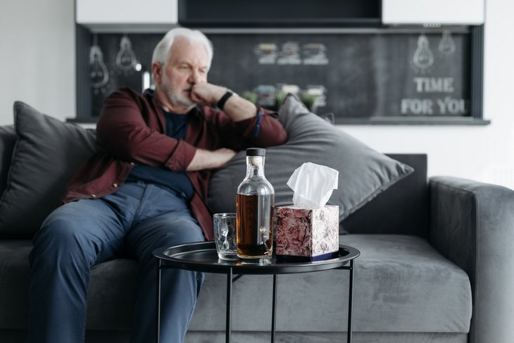 Na zdjęciu widoczny starszy mężczyzna siedzący na kanapie, wygląda na zadumanego. Na stoliku przed mężczyzną znajduje się butelka z alkoholem. Kadr szeroki. 