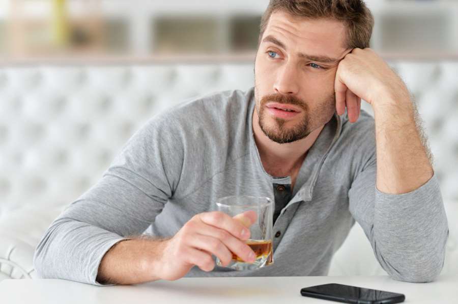 Na zdjęciu widoczny mężczyzna siedzący przy stole. Jest zamyślony, jedna ręką podpiera głowę w drugiej ręce trzyma szklankę z alkoholem. Kadr amerykański.