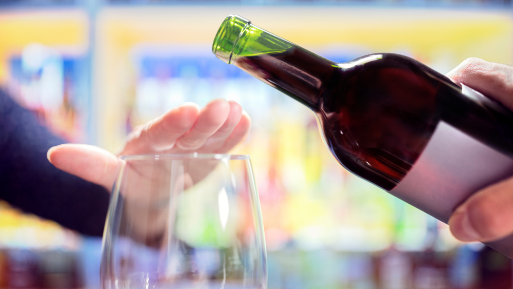 Na zdjęciu widoczny kieliszek oraz butelka wina. Dłoń zasłania kieliszek, co można rozumieć jako gest odmowy przed spożyciem alkoholu. Kadr wąski. 