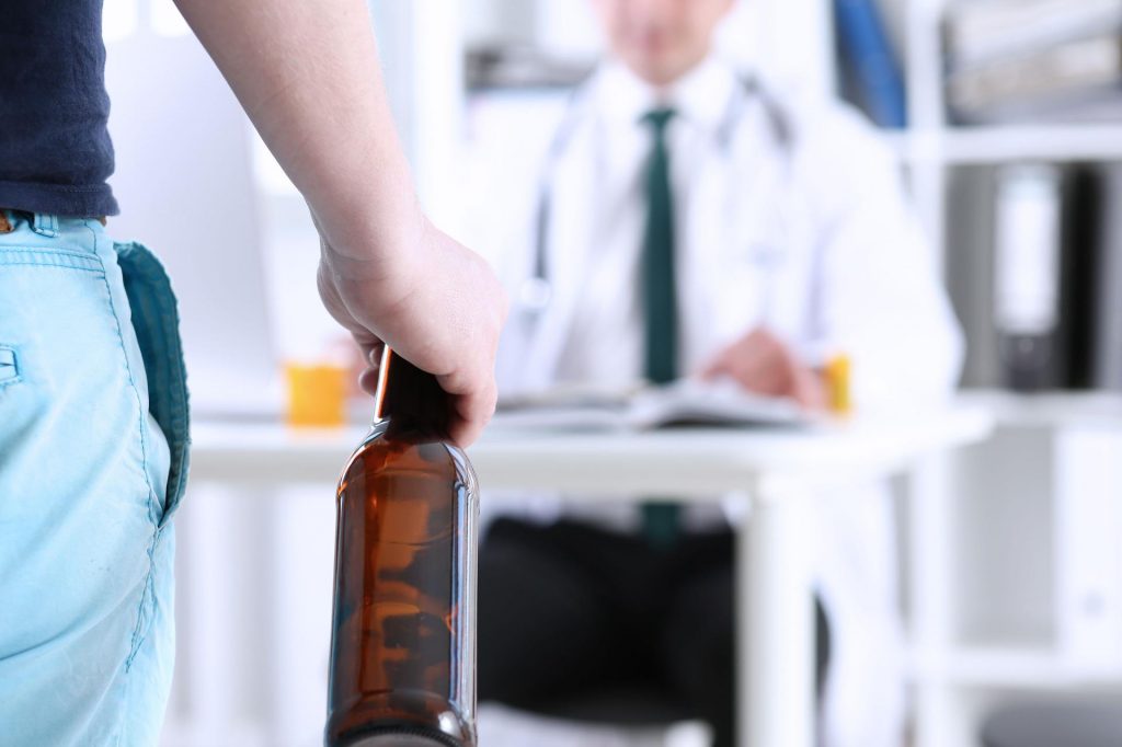 Na zdjęciu widoczny fragment sylwetki mężczyzny, który trzyma w ręku butelkę po alkoholu. Na drugim planie widoczna postać przypominająca lekarza. Kadr wąski. 