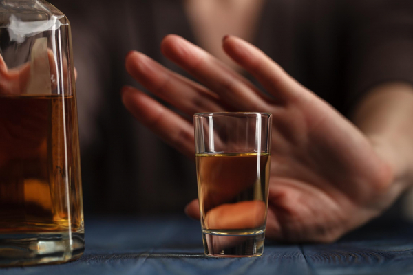 Na zdjęciu widoczny fragment butelki z alkoholem oraz szklanka napełniona alkoholem. Na drugim planie widoczna wyciągnięta w stronę szklanki ręka z dłonią ułożoną w geście sprzeciwu. Kadr wąski. 