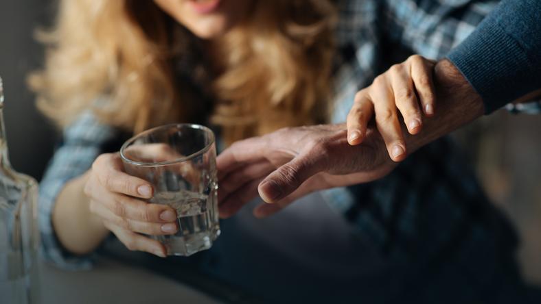 Na zdjęciu widoczna kobieca sylwetka, która jedną ręką przytrzymuje rękę mężczyzny a drugą zabiera z męskiej dłoni szklankę z alkoholem. Kadr wąski. 
