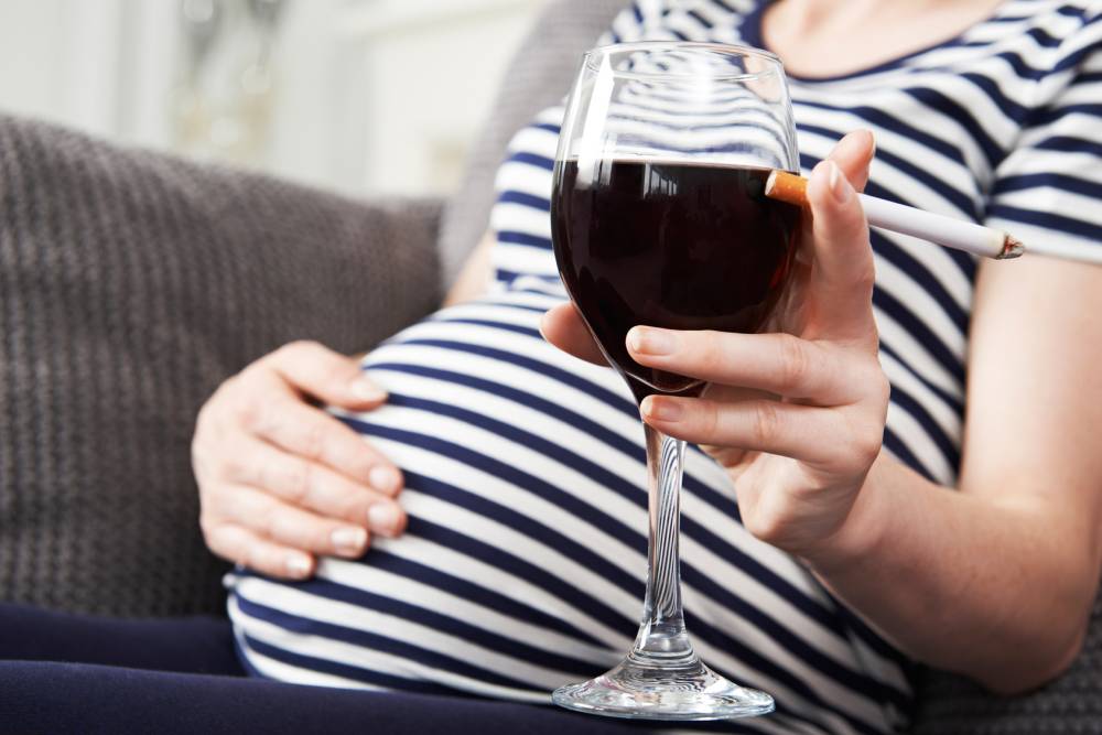 Na zdjęciu widoczny fragment sylwetki kobiety w ciąży siedzącej na kanapie, która w jednej z rąk trzyma kieliszek wina oraz zapalonego papierosa. Kadr wąski.