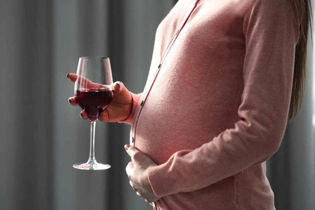 Na zdjęciu widoczny fragment kobiecej sylwetki z widocznym brzuchem ciążowym, która w jednej z dłoni trzyma kieliszek z winem. Kadr amerykański bez widocznej twarzy.