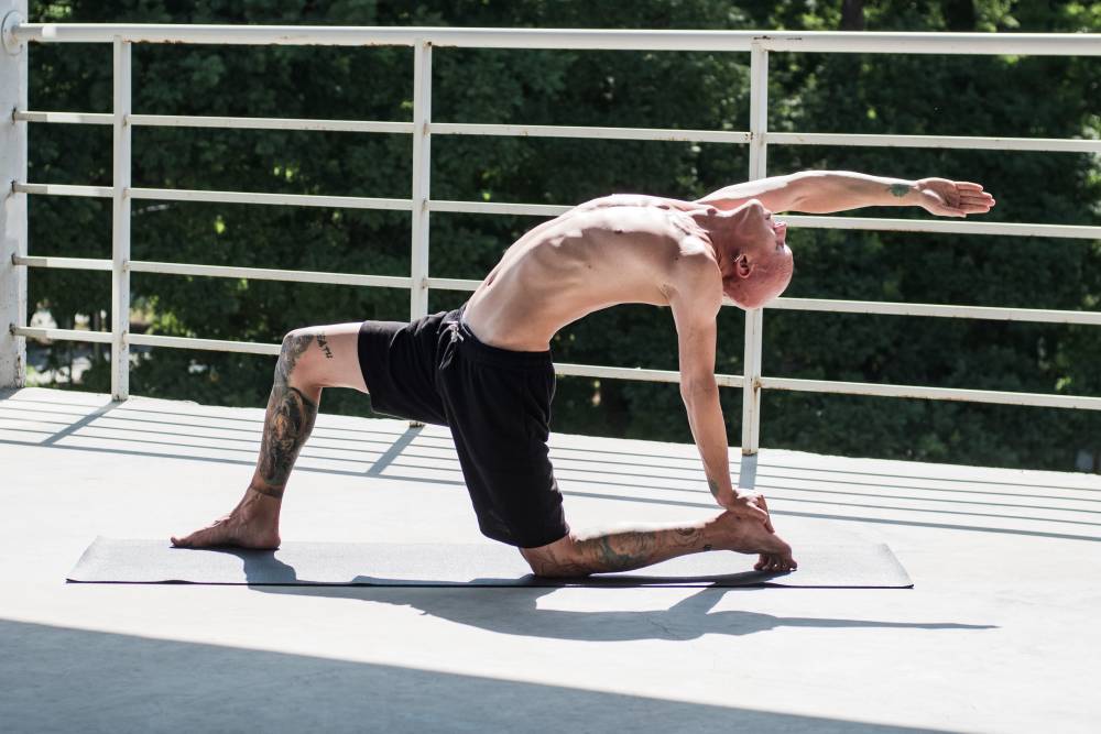 Na zdjęciu widoczny wytatuowany mężczyzna, który na tarasie ćwiczy jogę. Kadr szeroki. 
