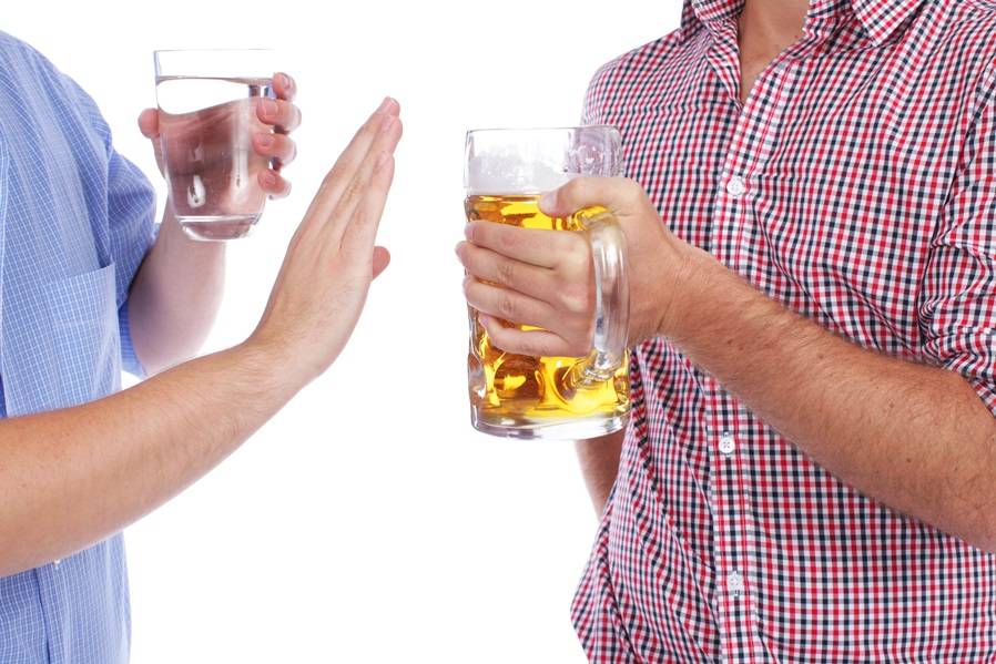 Na zdjęciu widoczne zarysy sylwetek dwóch mężczyzn. Jeden z nich, po lewej stronie, trzyma w rękach kufel z piwem, zachęcająco wysuwając go w stronę drugiego mężczyzny. Drugi mężczyzna z prawej strony w jednej ręce trzyma szklankę z wodą, a drugą rękę trzyma uniesioną w geście odmowy. Kadr wąski. 