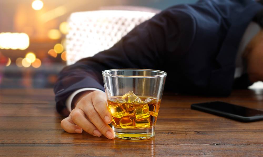 Na zdjęciu widoczna sylwetka mężczyzny trzymającego w dłoni szklankę z alkoholem. Mężczyzna prawdopodobnie zasną oparty o stolik. Kadr wąski. 