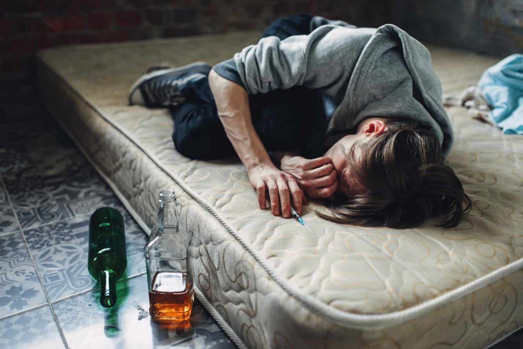 Na zdjęciu widoczny młody mężczyzna śpiący na brudnym materacu, wokół którego porozrzucane są butelki po alkoholu. Kadr szeroki. 