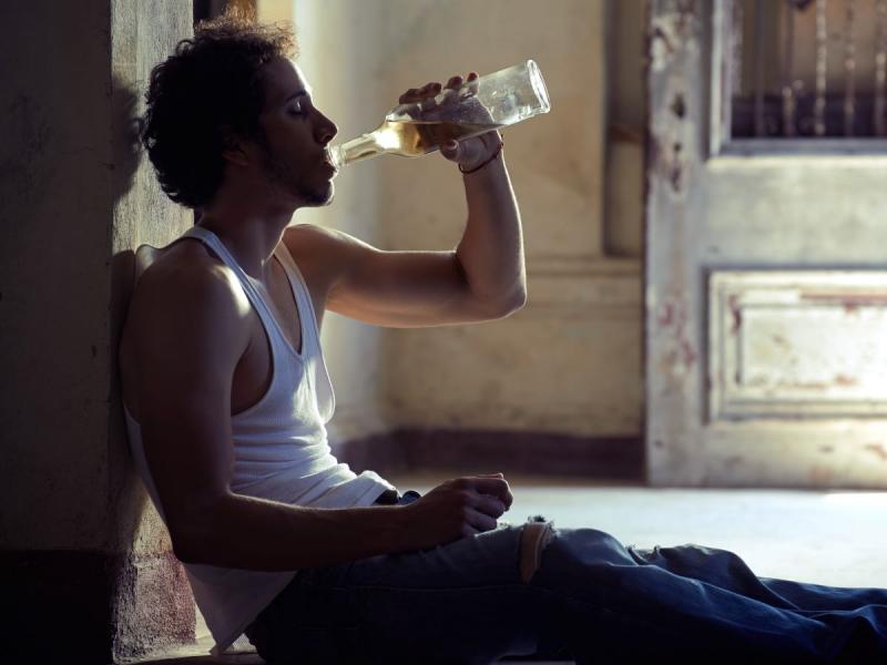 Na zdjęciu widoczny mężczyzna siedzący na podłodze i oparty plecami o ścianę. Mężczyzna spożywa alkohol prosto z butelki. Kadr szeroki. 