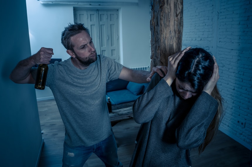 Na zdjęciu widoczny mężczyzna oraz kobieta. Mężczyzna w jednej ręce trzyma butelkę alkoholu i przejawia agresywne zachowania wobec kobiety. Kadr amerykański. 