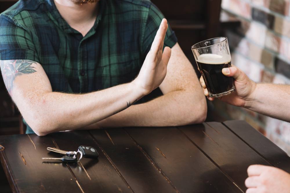 Na zdjęciu widoczna sylwetka mężczyzny bez pokazanej twarzy, oraz ręką serwująca alkohol. Mężczyzna gestem odmawia przyjęcia alkoholu. Kadr wąski. 
