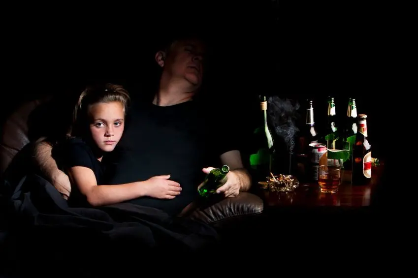 Na zdjęciu widoczny mężczyzna – ojciec śpiący na siedząco na kanapie. Obok na stoliku znajdują się liczne butelki po alkoholu. W mężczyznę wtulona jest mała dziewczynka – córka, ze smutnym wyrazem twarzy. Kadr szeroki. 