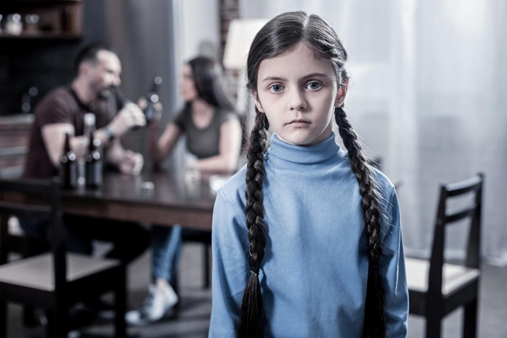 Na zdjęciu na pierwszym planie widoczna mała dziewczyna ze smutnym wyrazem twarzy. Na drugim planie widać stół, przy którym siedzą rodzice pijący alkohol. Kadr szeroki. 