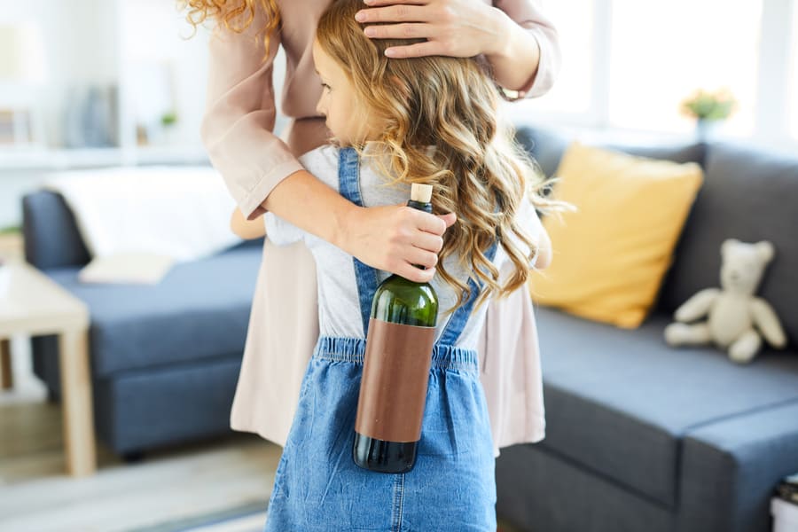 Na zdjęciu widoczna mała dziewczynka tyłem przytulająca się do swojej mamy. Mama w jednej ręce trzyma butelkę wina. Kadr amerykański. 
