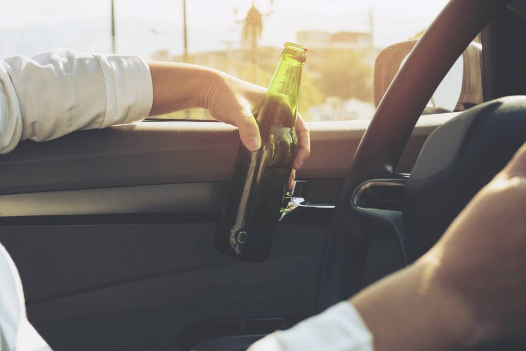 Na zdjęciu widoczne wnętrze samochodu od strony kierowcy wraz z zarysem męskiej sylwetki kierującej pojazdem. Kierujący trzyma w ręce butelkę piwa. Kadr wąski. 