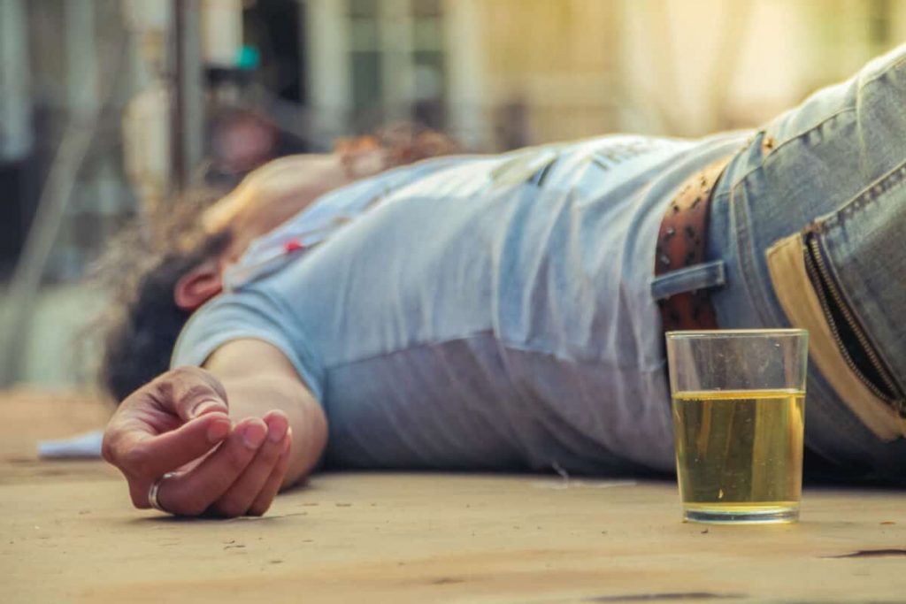 Na zdjęciu widoczna postać mężczyzny leżącego na podłodze, na pierwszym planie widać również szklankę z alkoholem. Kadr szeroki. 