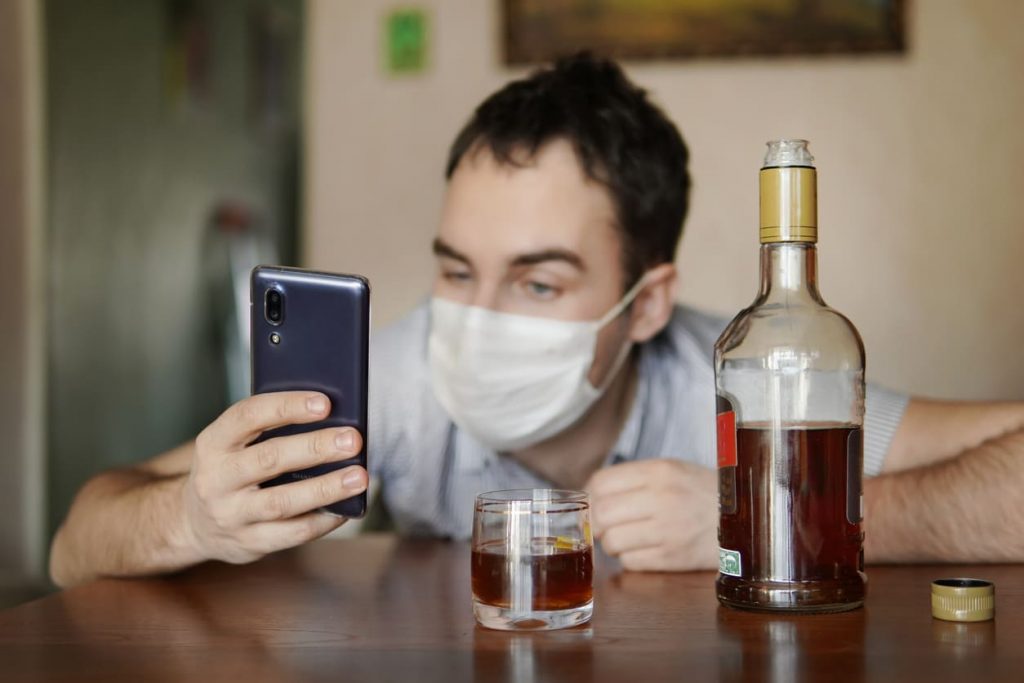 Na zdjęciu widoczny mężczyzna siedzący przy stole, na którym stoi butelka alkoholu oraz szklanka z nalanym alkoholem. Mężczyzna ma na twarzy maseczkę medyczną. Kadr amerykański.