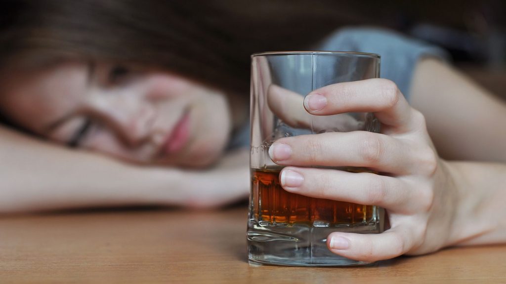 Na pierwszym planie zdjęcia widoczna szklanka z whisky, z zaciśnięta na niej kobieca dłoniach. Na drugim planie widoczna nastolatka, która przysypia oparta o ramię na stole. Kadr szeroki. 