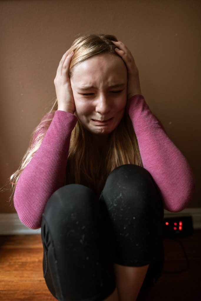 Na zdjęciu widoczna młoda kobieta siedząca na podłodze z podkurczonymi nogami – zatyka uszy rękami i płacze. Kadr amerykański. 