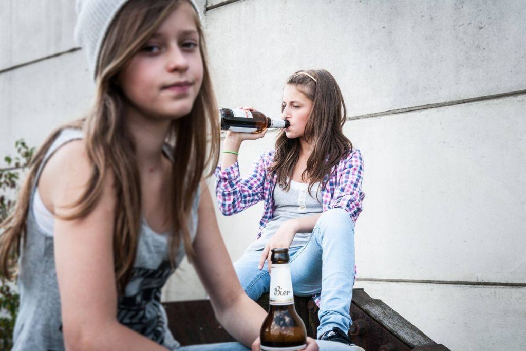 Na zdjęciu widoczne dwie nastoletnie dziewczyny. Na pierwszym planie siedząca nastolatka trzyma w ręku butelkę piwa. Na drugim planie siedząca luźno nastolatka pije piwo. Kadr szeroki, ostrość na drugi plan. 