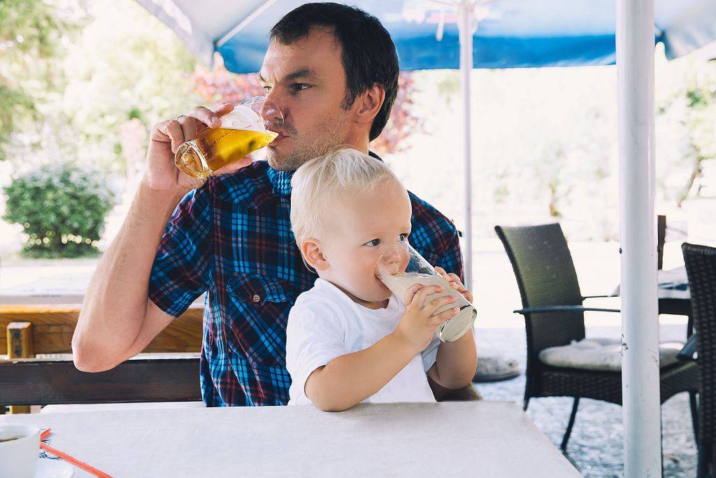 Na zdjęciu widoczny mężczyzna trzymający na kolanach małe dziecko. Mężczyzna pije piwo, dziecko inny napój. Kadr szeroki. 
