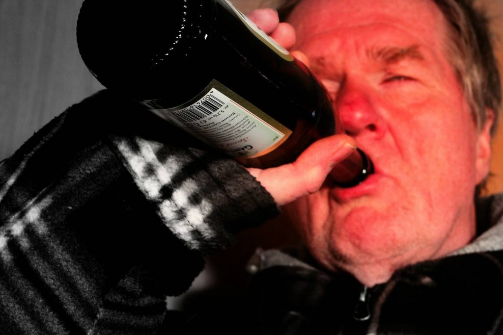 Na zdjęciu widoczny mężczyzna pijący piwo z butelki. Jego twarz jest zaczerwieniona. Kadr portretowy z ostrością na pierwszy plan. 