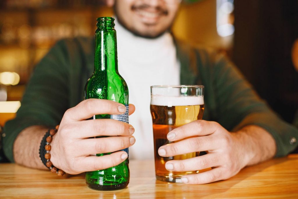 Na zdjęciu widoczny mężczyzna, trzymający w ręku piwo oraz szklankę do piwa. Ostrość na pierwszy plan, kadr amerykański bez pokazanej twarzy. 