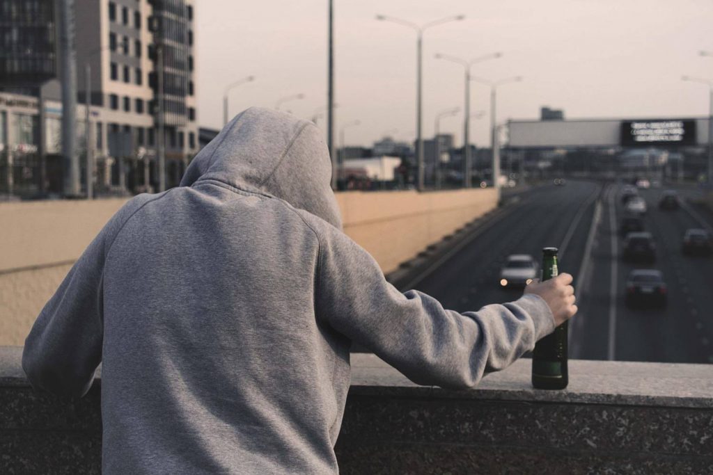 Na zdjęciu widoczny mężczyzna stojący na moście z zarzuconym na głowę kapturem, w ręku trzyma butelkę piwa. Kadr amerykański, ujęcie tyłem. 
