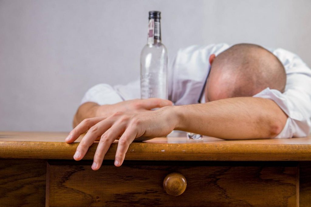 Na zdjęciu widoczny mężczyzna z opartą na przedramieniu głową, na pierwszym planie widoczna butelka po wypitym alkoholu. Kadr portretowy. 