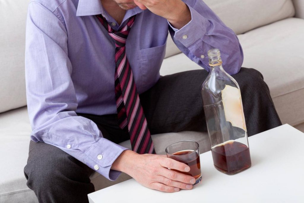 Na zdjęciu widoczny elegancko ubrany mężczyzna siedzący przy stoliku i trzymający w ręku szklankę z whisky. Kadr centralny, bez widocznej twarzy. 