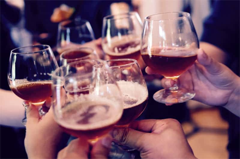 Na zdjęciu widoczne ręce wielu osób, które „stukają się” szklankami i kieliszkami zawierającymi różne alkohole. Kadr centralny. 