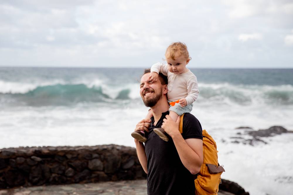 Na zdjęciu widoczny szczęśliwy mężczyzna niosący małe dziecko na barana. W tle widoczne plaża oraz może. Kadr amerykański. 
