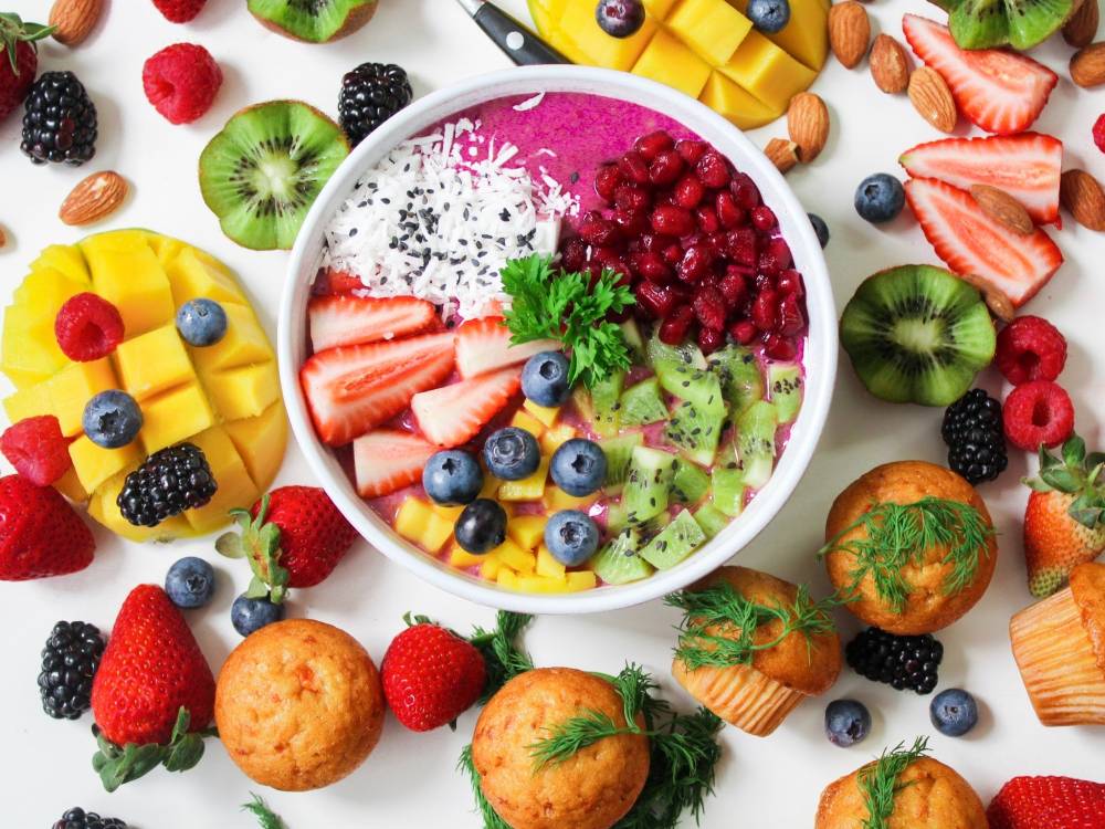 Na zdjęciu widoczne różnokolorowe warzywa, owoce, pieczywo. W centrum znajduje się miska z owsianką oraz różnymi owocami. Kadr centralny. 
