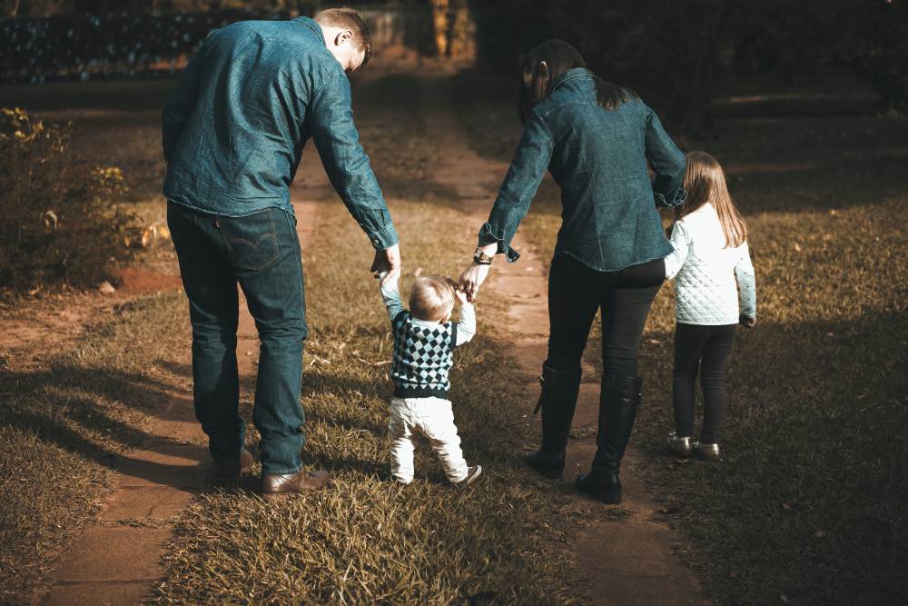 Na zdjęciu widoczna rodzina – kobieta, mężczyzna oraz dwójka małych dzieci idących z rodzicami za rękę po leśnej ścieżce. Kadr od tyłu. 