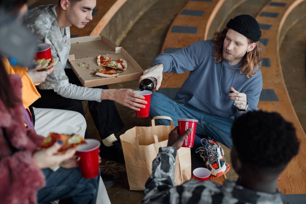 Na zdjęciu widoczna grupa osób jedząca pizzę. Jeden z mężczyzn polewa innym alkohol w plastikowe kubki. Kadr szeroki. 