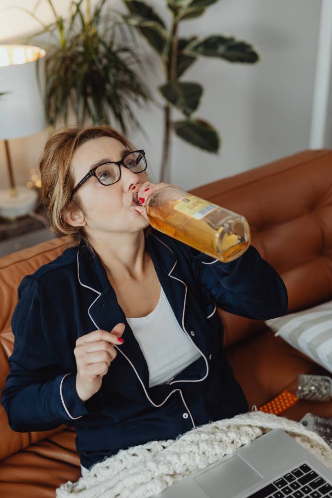 Na zdjęciu widoczna kobieta siedząca na kanapie i prawdopodobnie pracująca wcześniej na laptopie. Kobieta pije prosto z butelki whisky. Kadr amerykański.  