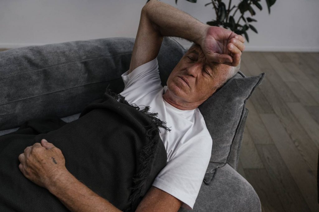 Na zdjęciu widoczny jest starszy mężczyzna, który przykryty kocem leży na sofie. Jego prawa dłoń spoczywa na czole. Widać, że osoba na zdjęciu źle się czuje. Kadr szeroki. 