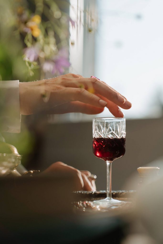 Na zdjęciu widoczny kieliszek napełniony czerwonym winem, stający na stoliku oraz dłoń. Palec dłoni oparty jest o krawędź kieliszka. Kadr portretowy. 