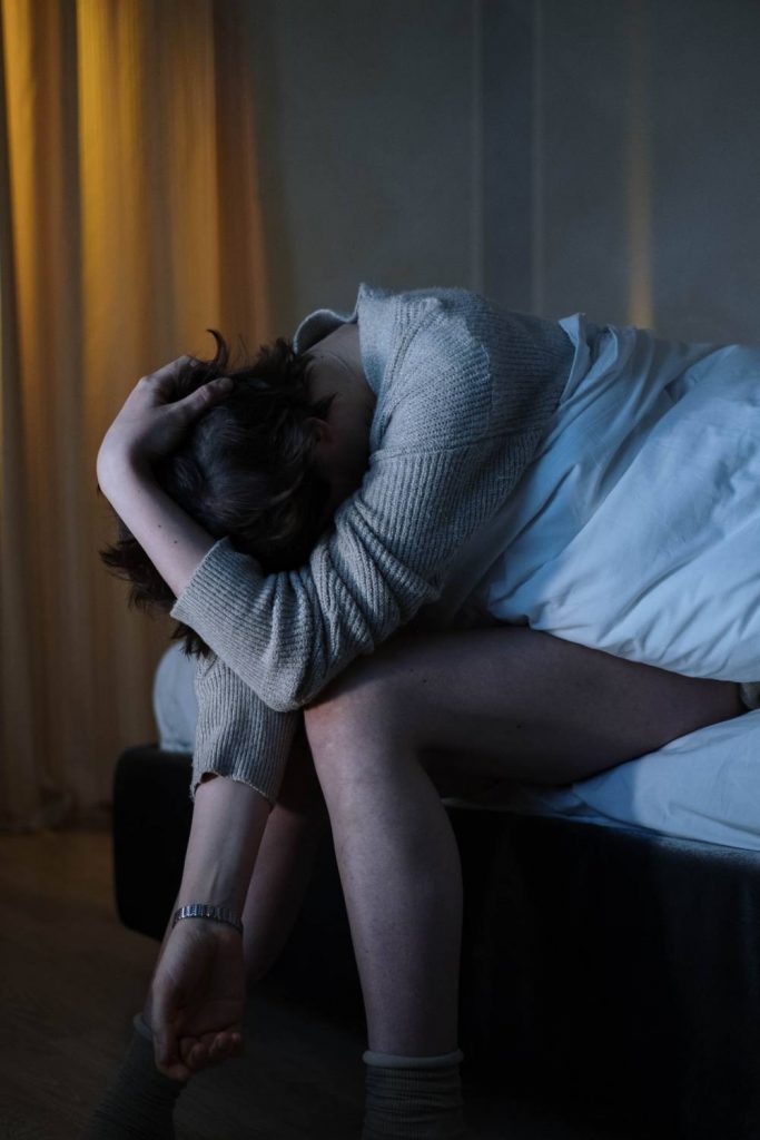 Na zdjęciu widoczna kobieta siedząca na łóżku, z rękami opartymi na kolanach w których skrywa głowę, sprawia wrażenie, że czuje się źle. Kadr amerykański. 