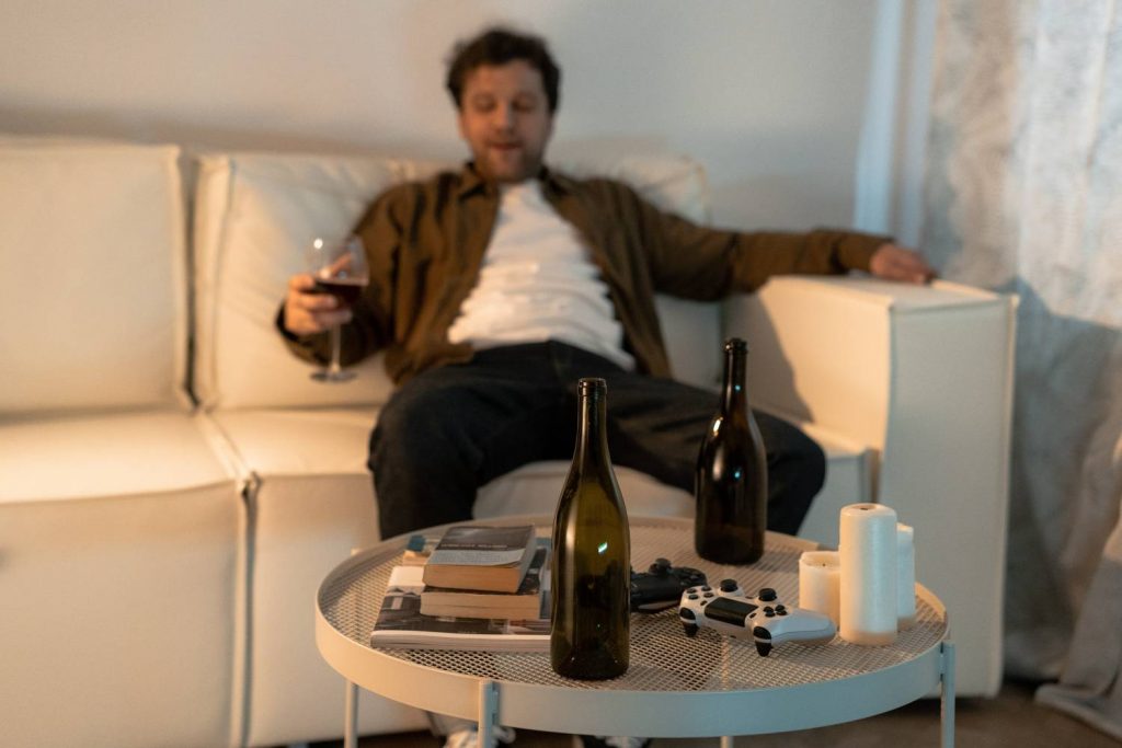 Zdjęcie przedstawia mężczyznę siedzącego na kanapie, w prawej ręce trzyma on kieliszek z winem. Postać widoczna na drugim planie. Na pierwszym planie widoczny stolik na którym znajdują się między innymi 2 butelki po winie. 