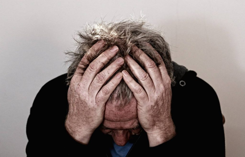 Zdjęcie przedstawia starszego mężczyznę z pochylona głową schowana w dłoniach. Zdjęcie emanuje smutnymi emocjami. Kadr portretowy. 