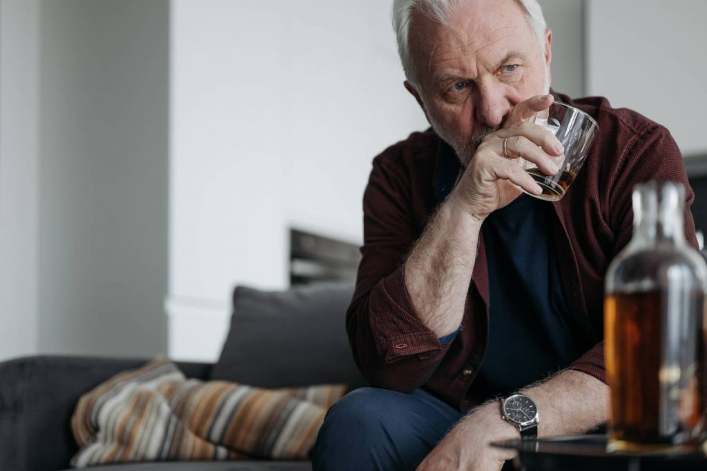 Zdjęcie przedstawiające starszego mężczyznę siedzącego na kanapie. W prawej ręce trzyma on szklankę do whisky. Na pierwszym planie po lewej stronie zdjęcia znajduje się stolik z karafką z whisky. Kadr amerykański. 